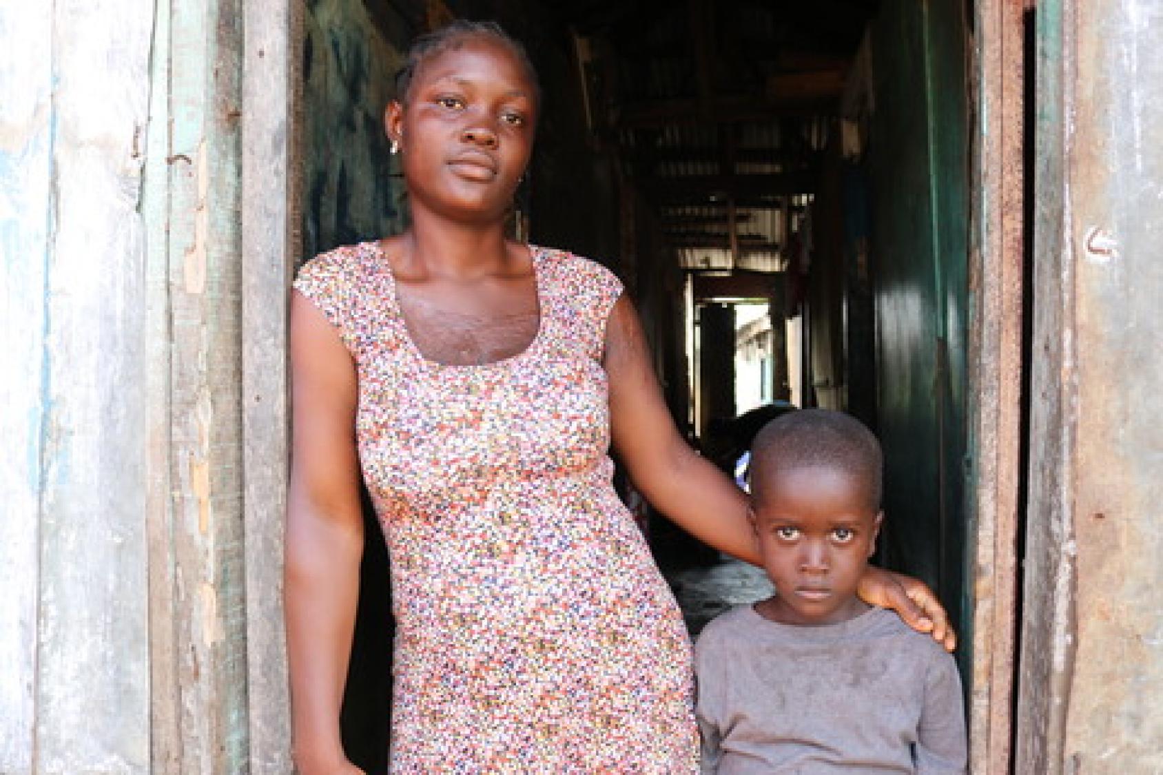 Молодая либерийская женщина стоит на пороге своего дома, обняв за плечи своего маленького сына.