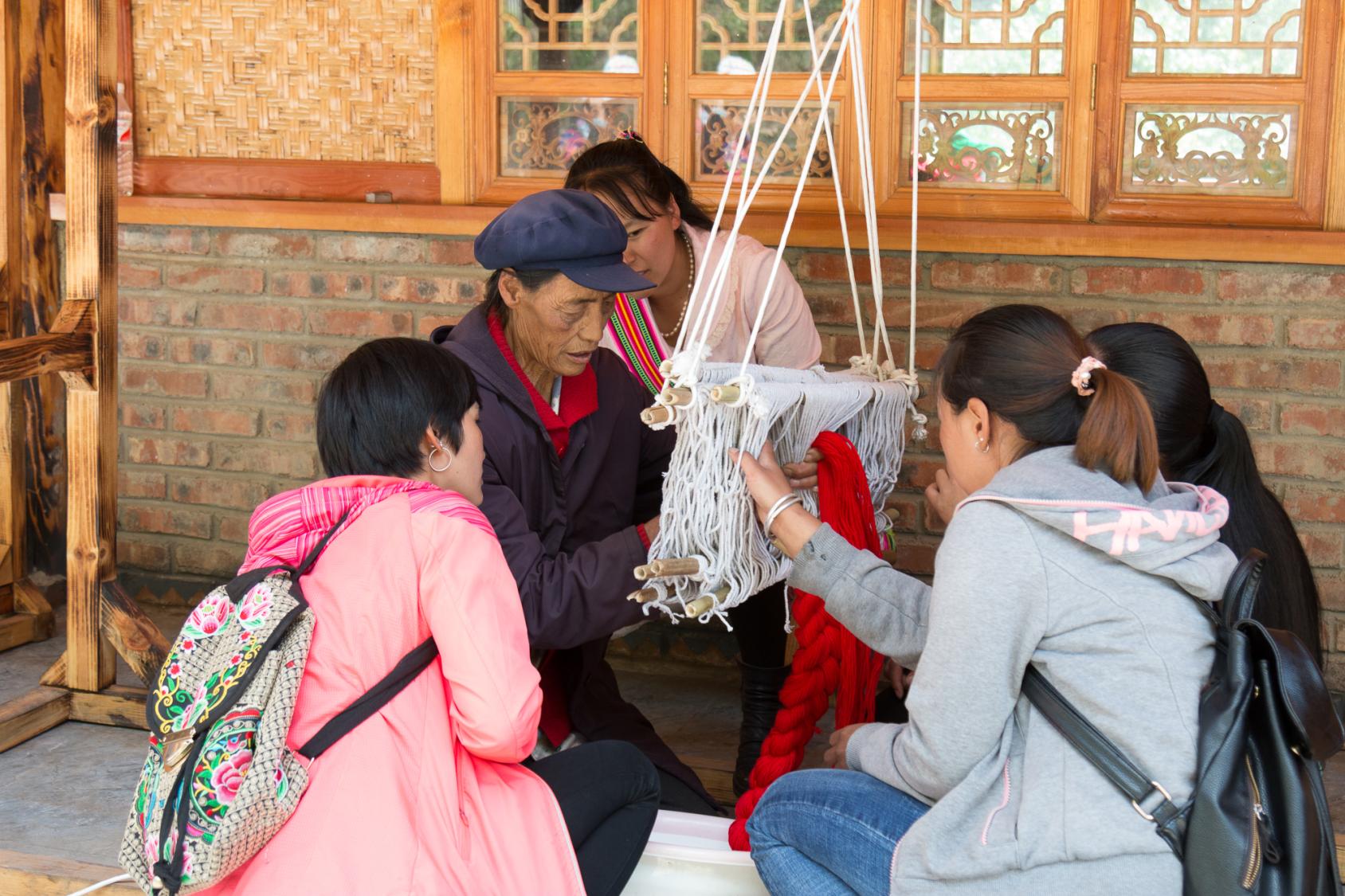 4 young women sitting around an old woman, watching her weave wool 