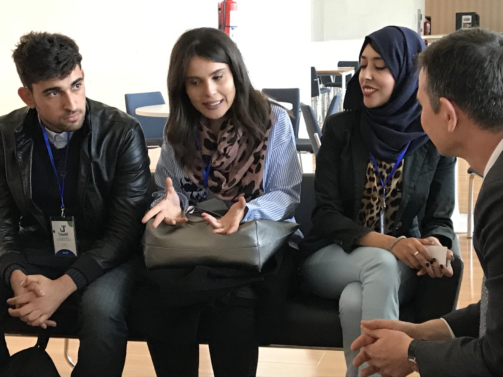 Two female graduates - one of whom wears a head scarf - and two male graduates sit in a training room and talk. They each wear a badge around their neck.
