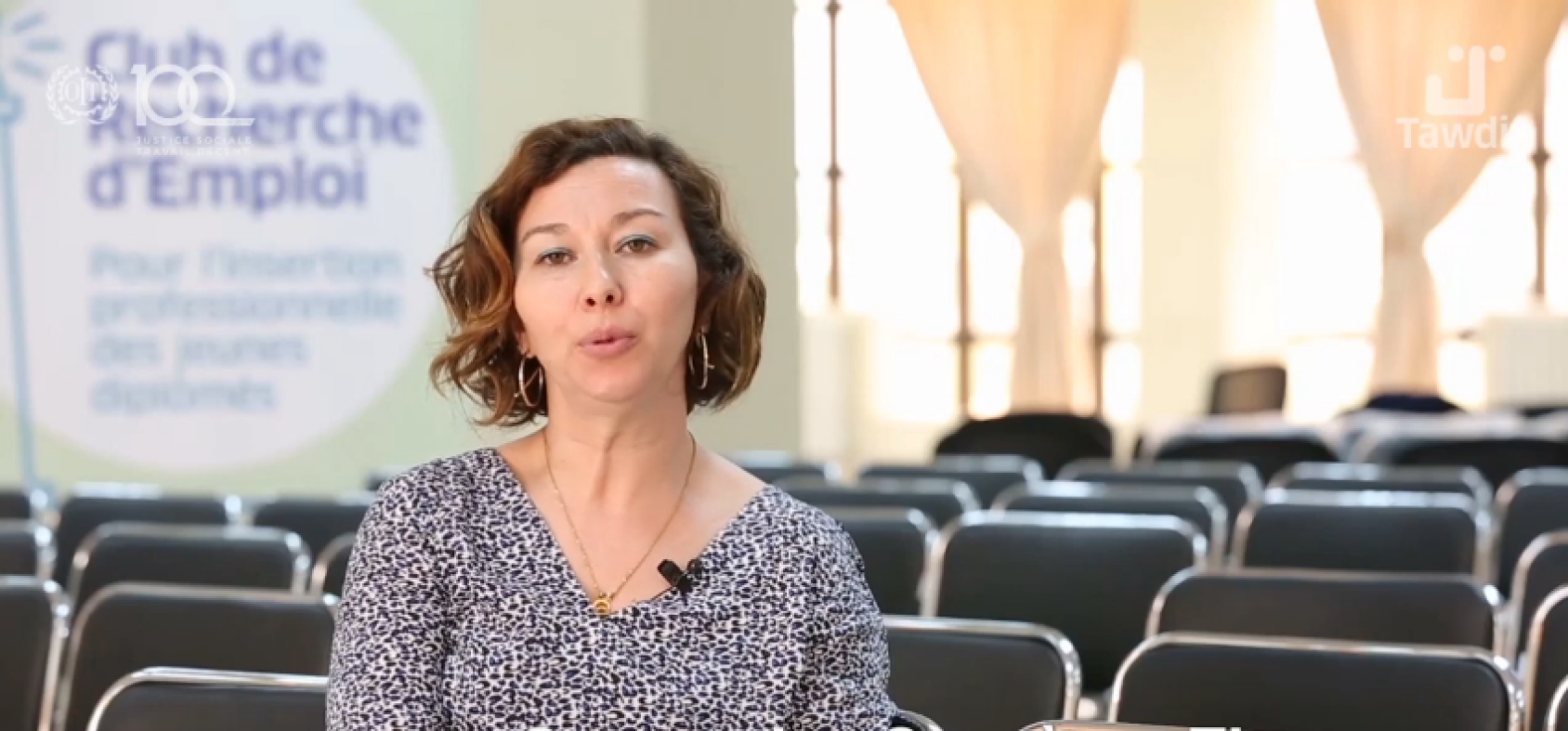 A woman with short hair sits in a conference room and speaks to the camera. In the background, on the left of the image, one can read the "Club de Recherche d'Emploi", which means "Job Search Club".