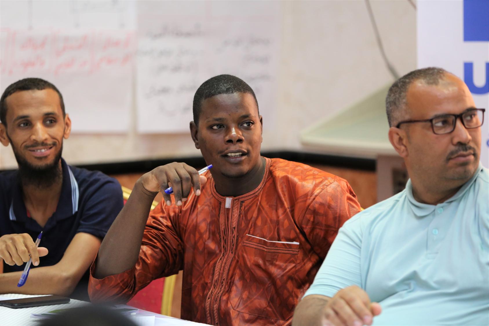 Three men are sitting side by side in a training room, looking at a person out of the camera's reach.