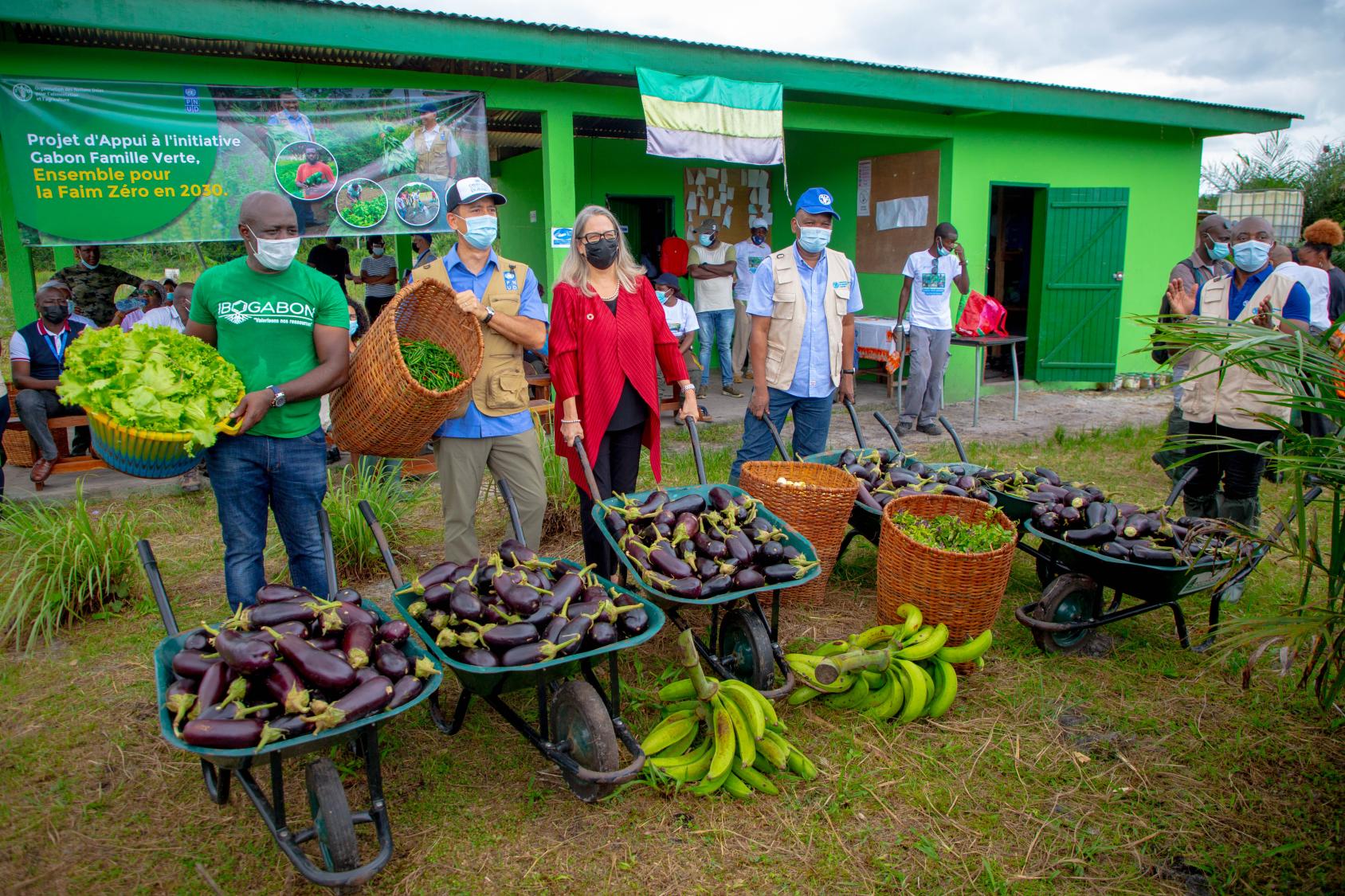 People at a small market for fruits and vegetables, in front of a green-colored building.