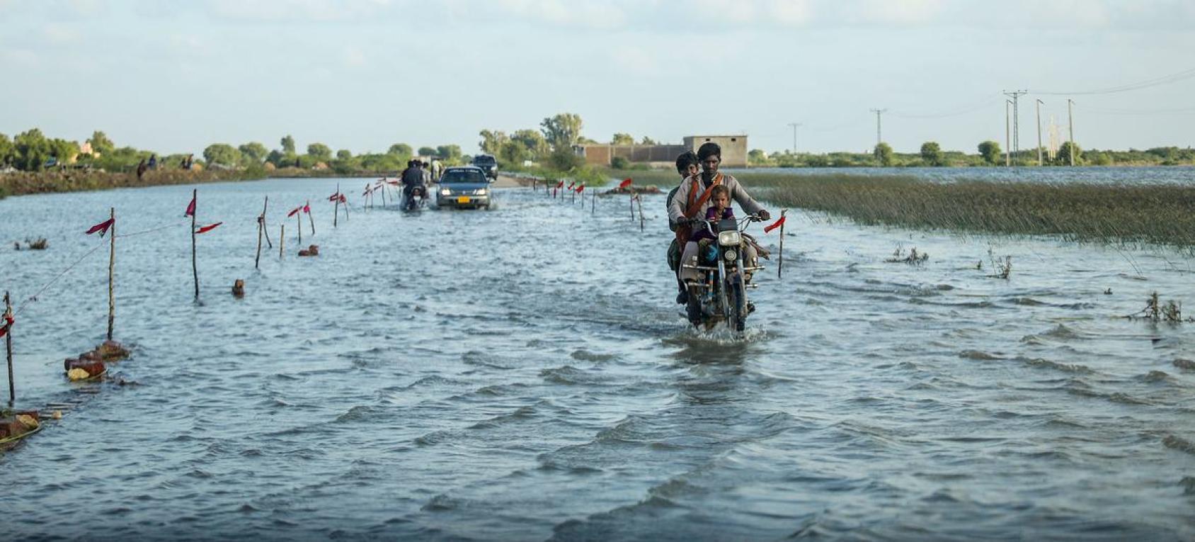 Two men are riding on a bike through a flooded area.