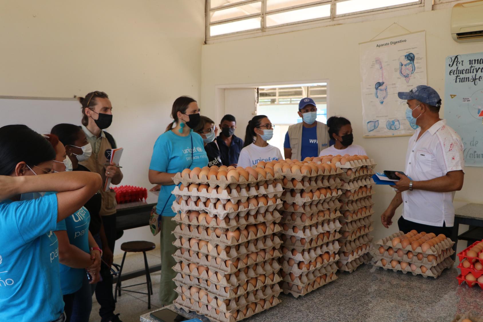 Un grupo de personas con mascarillas conversan en la parte interna de una edificación escolar, y en el centro hay tres torres de cartones de huevos.