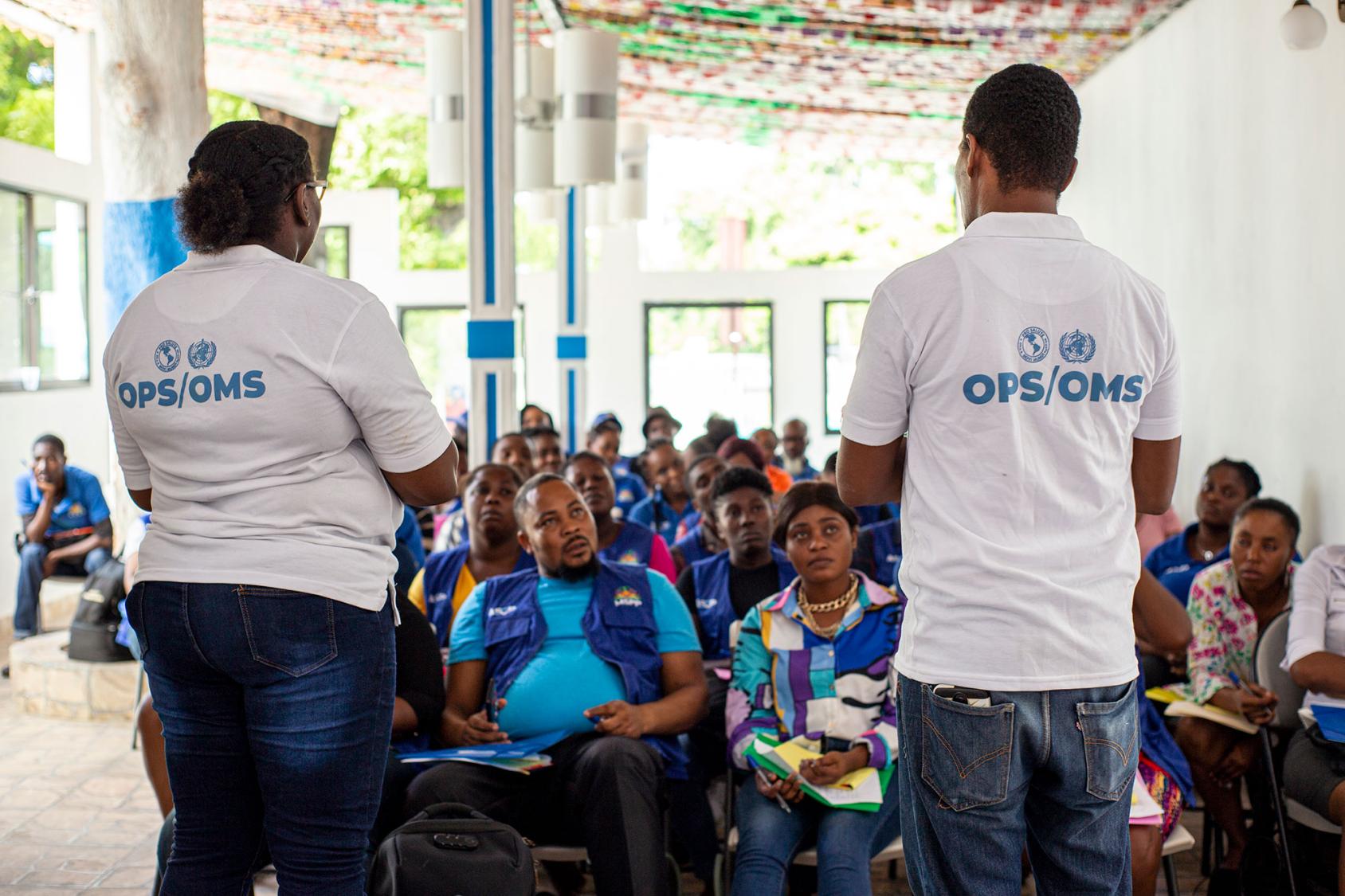Two PAHO/WHO employees, both wearing jeans and white t-shirts, speak to community health workers in a training room.