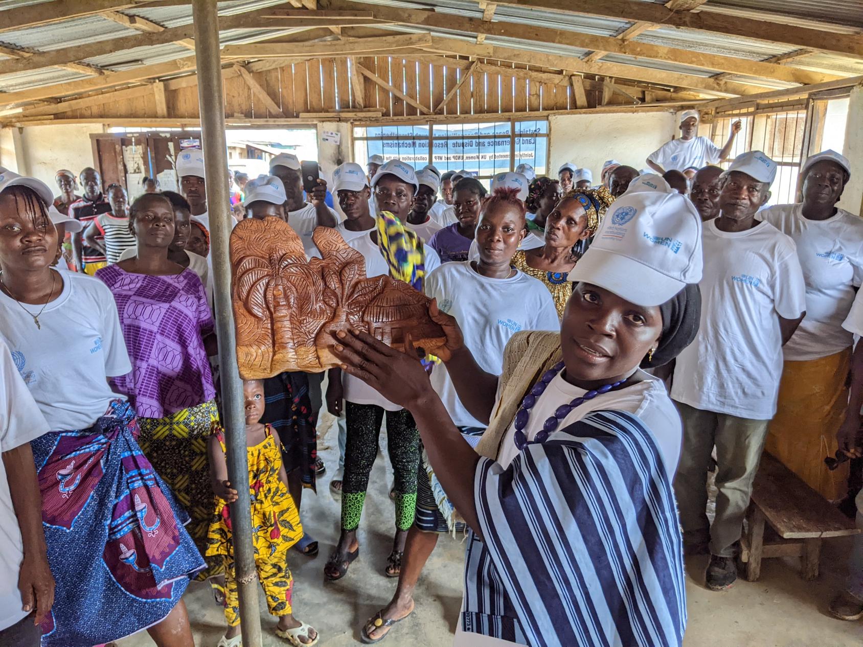 In Liberia, a group of women and men are gathered in a room and look towards the camera as one of the women, in the foreground, shows a handmade sculpture. 