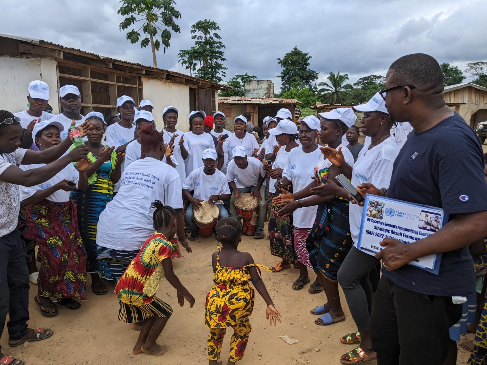 A woman and two girls dance in the middle of a ring of people wearing white shirts and caps, to the rhythm of drums played by two men.