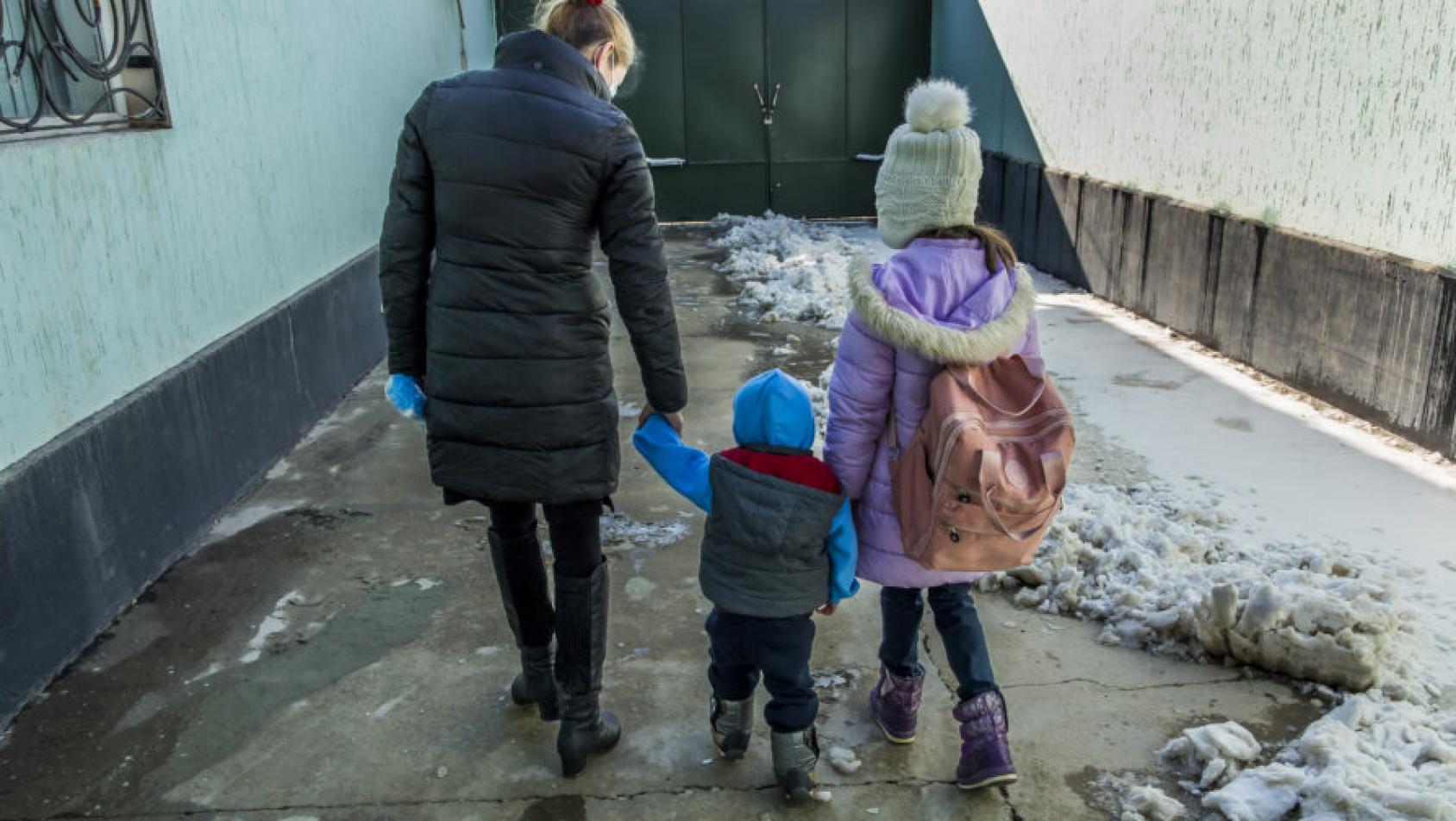 En un pasillo exterior, con nieve en un lateral, se ve de espaldas a una mujer caminando de la mano con dos pequeños, y todos van con ropa de abrigo.
