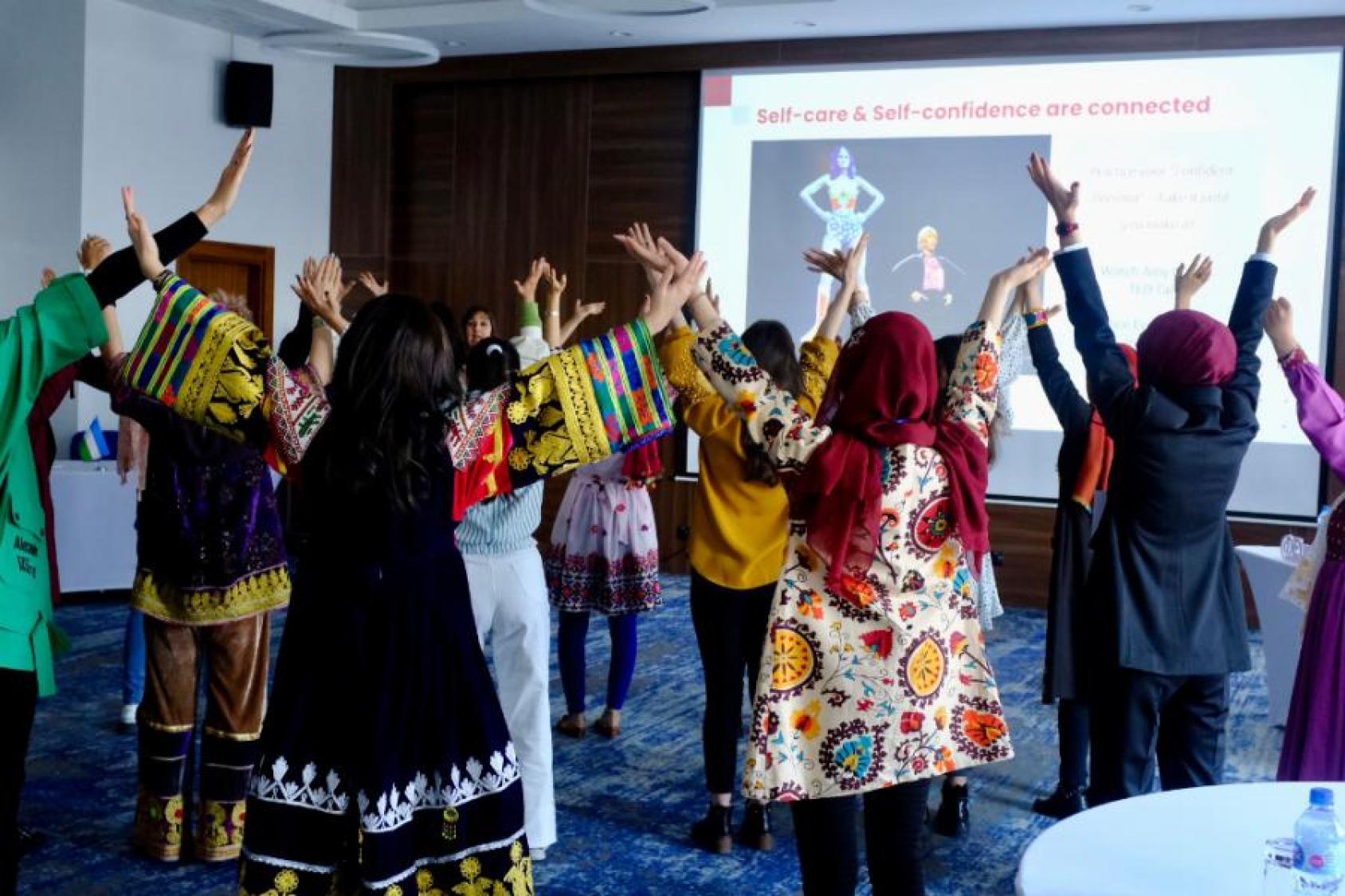 A small group of females stand in a circle and stretch with their arms raised.