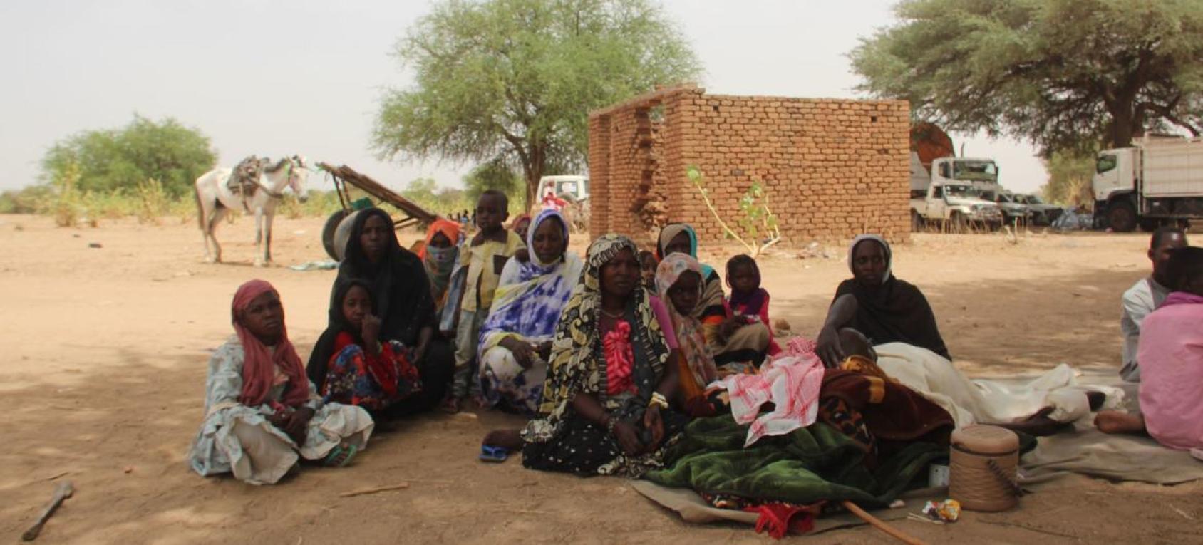 Group of people in different coloured clothes sitting in a barren rural field.