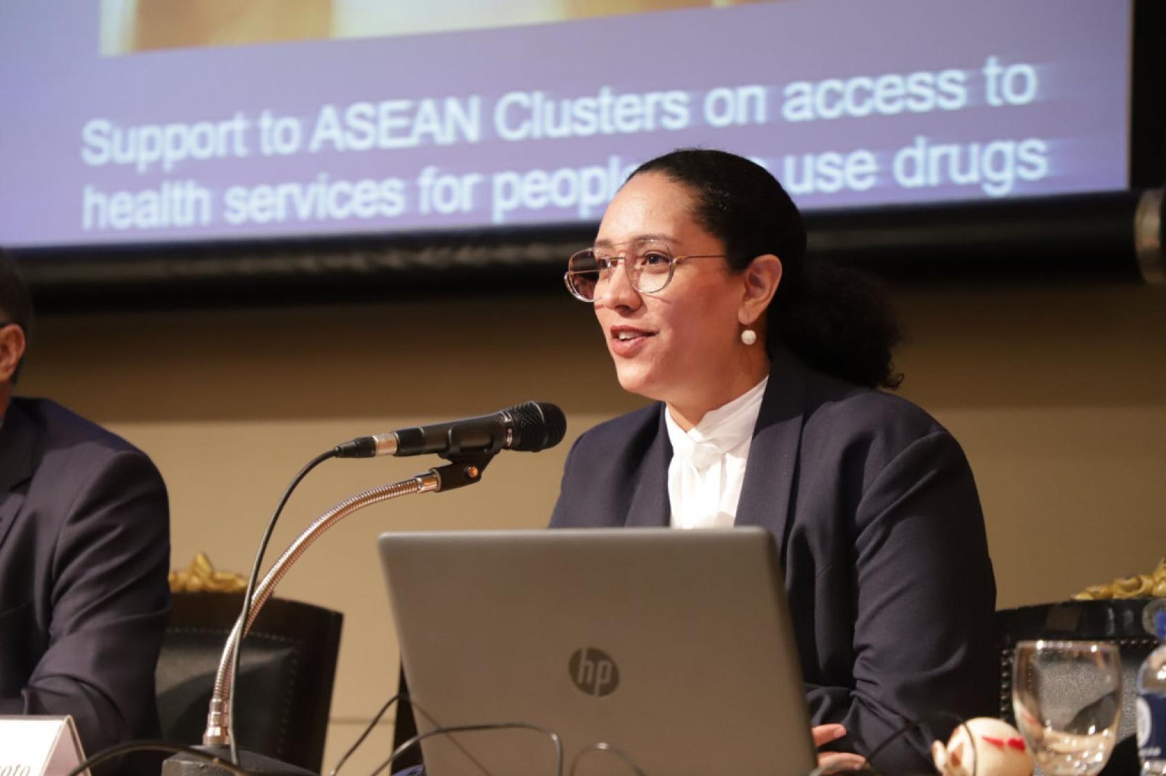 A woman in a black suit and spectacles sits at a podium and speak into a microphone