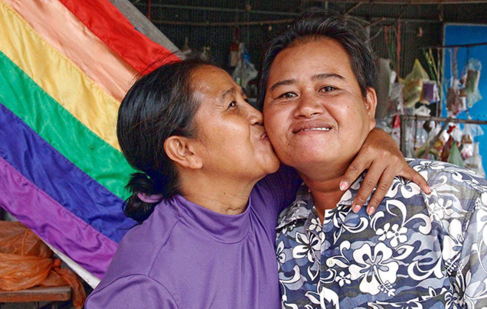A person in a purple shirt kisses another person in a printed shirt on the cheek in front of a rainbow coloured flag