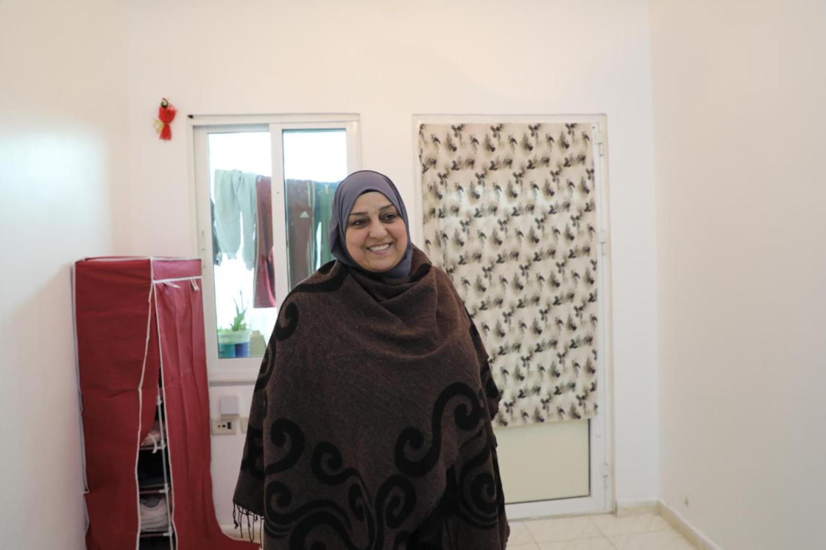 Woman in a dark brown dress and headscarf stands in a room in front of. window