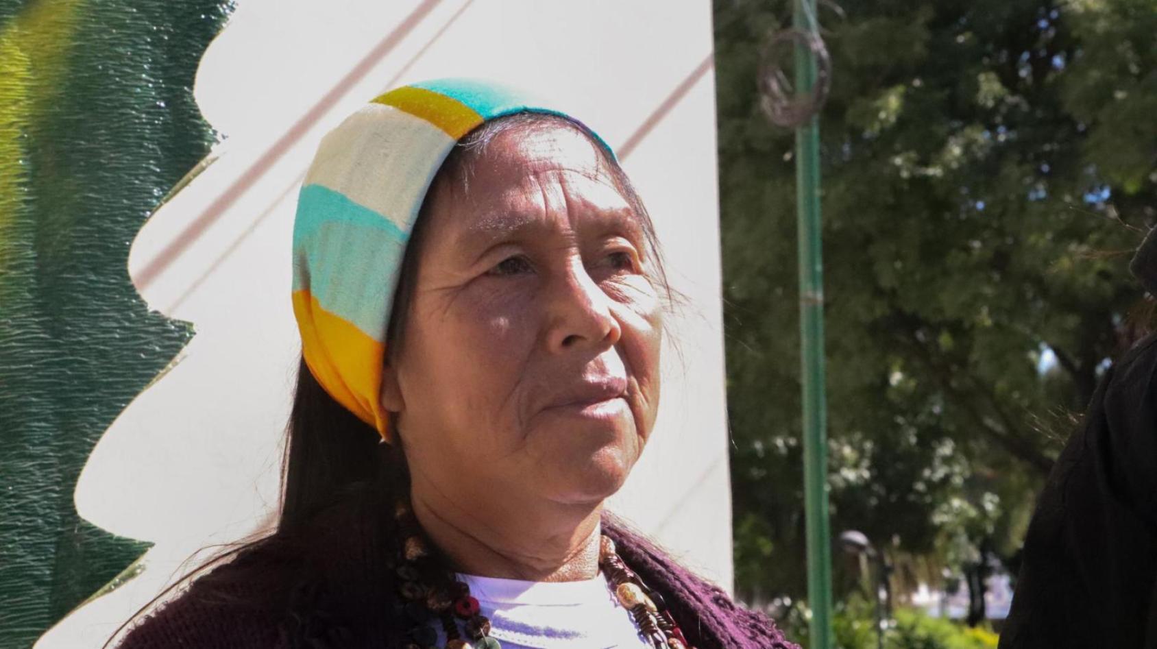 An indigenous woman in a green and orange headband stands in front of a greenery-filled background