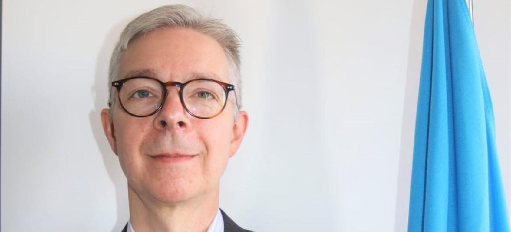 A UN official, in a suit and glasses, stands next to a UN flag