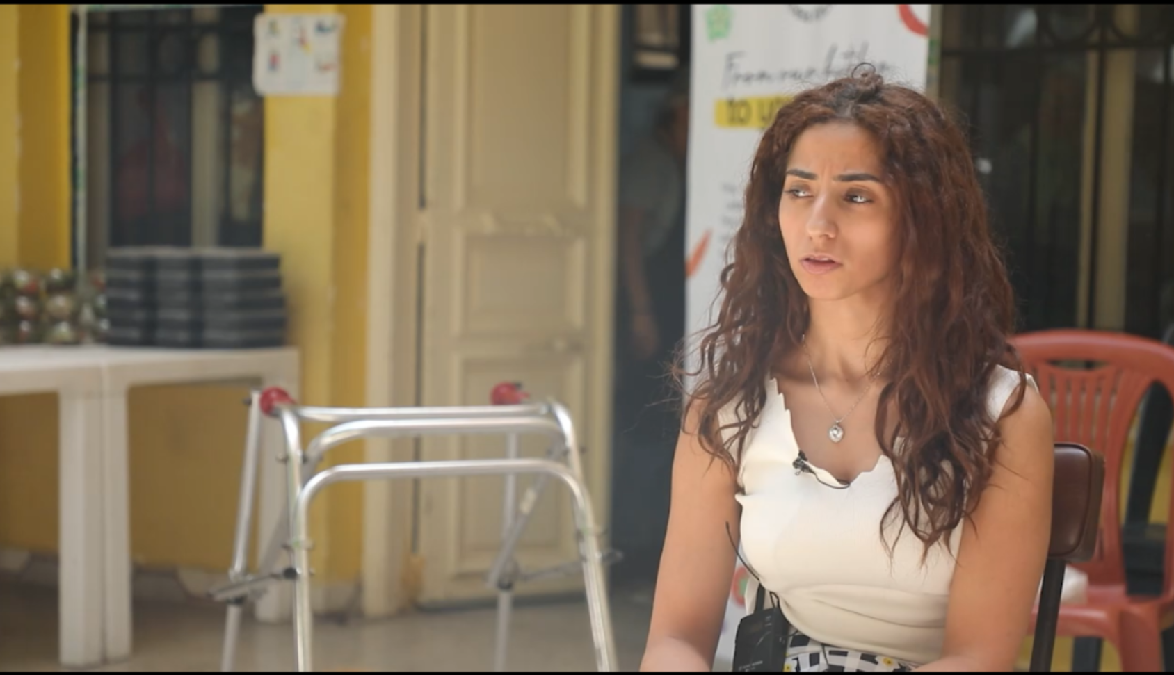 A woman with curly hair in a white tank top talks and faces the camera.