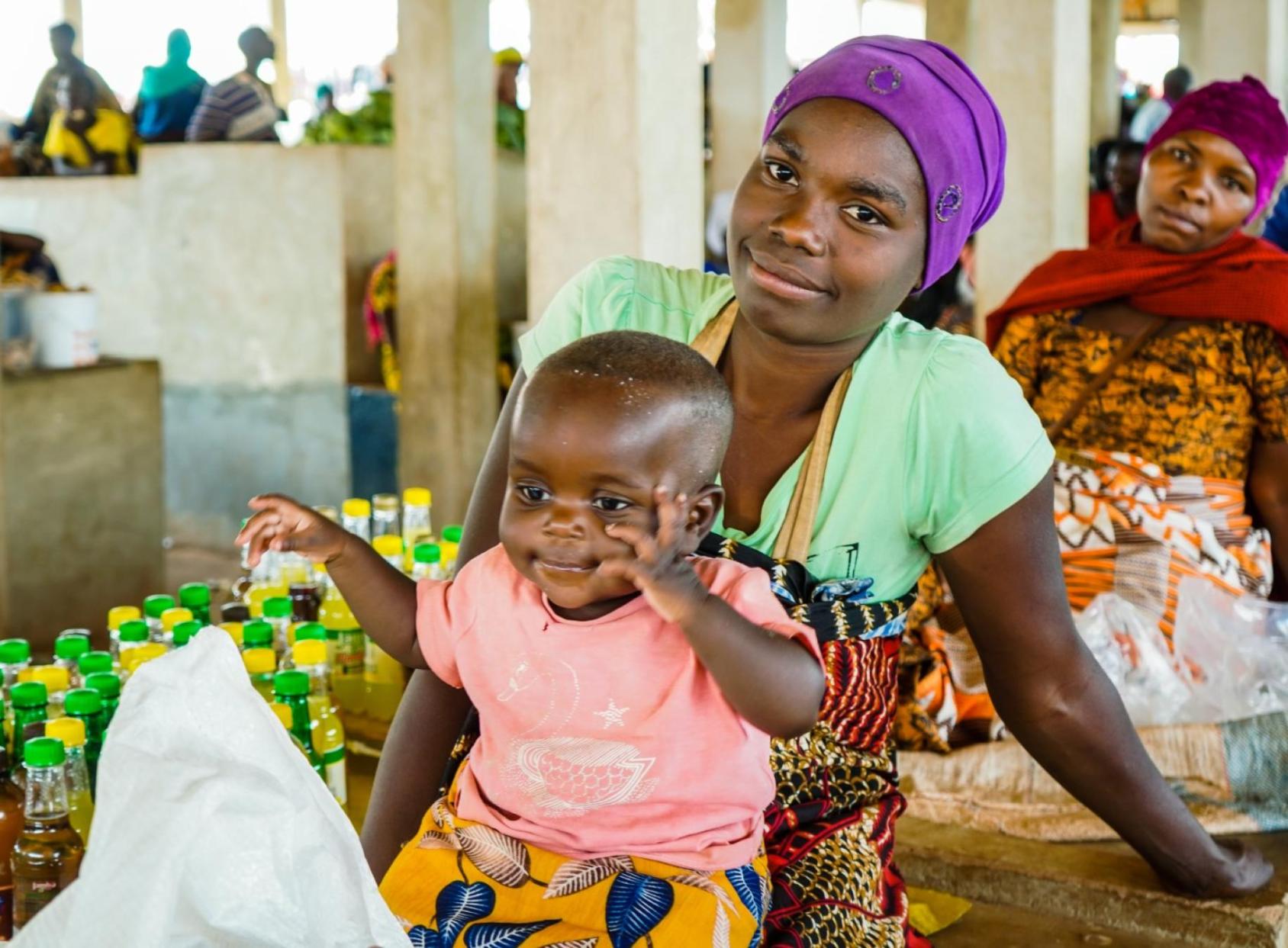 Girl in a green dress and purple headscarf holds a small baby in a pink dress in a market place