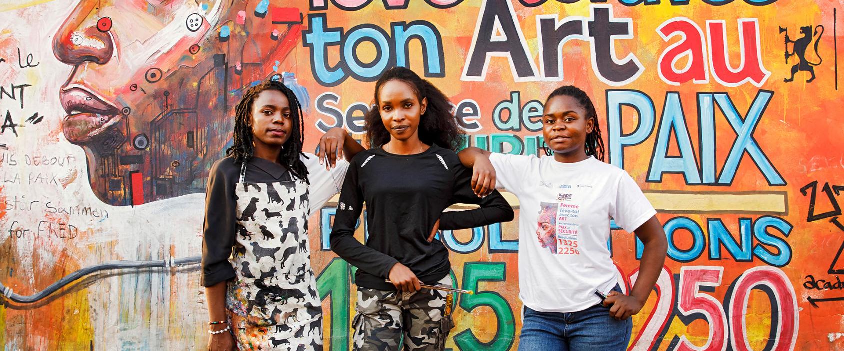 Three young women standing in front of a painted mural depicting words of peace