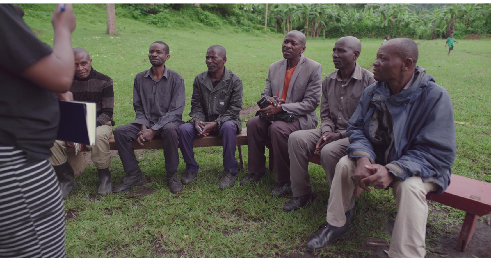 A group of Ugandan men sit around a bench while one of them seems to be speaking to them.