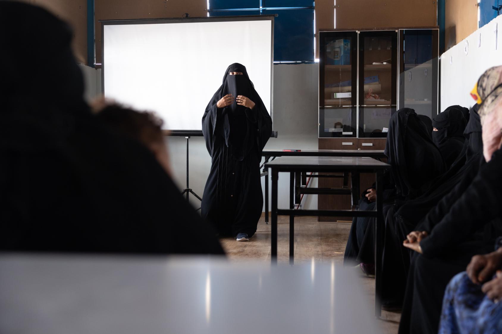 A woman in a black abaya, fully covered from head to toe, stands in front of a room, teaching other women.