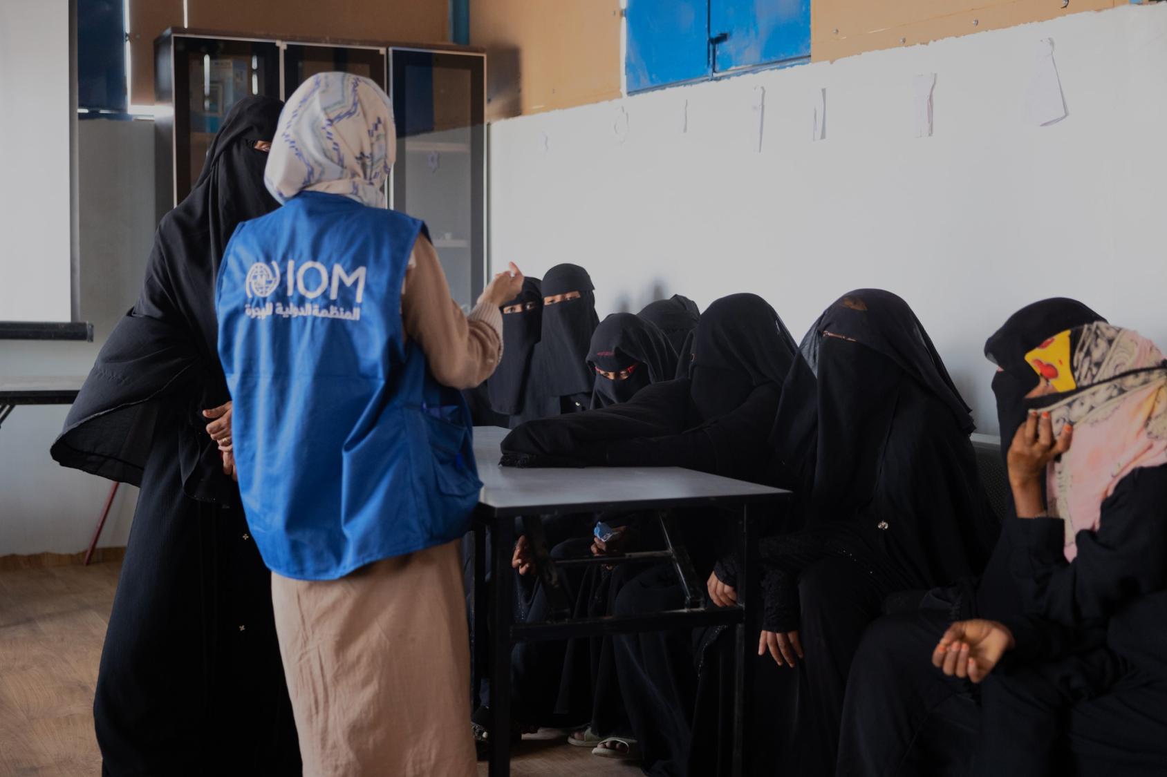 A woman in a blue vest with the IOM logo stands in front of a desk where several women and girls, fully clothed in black listen.