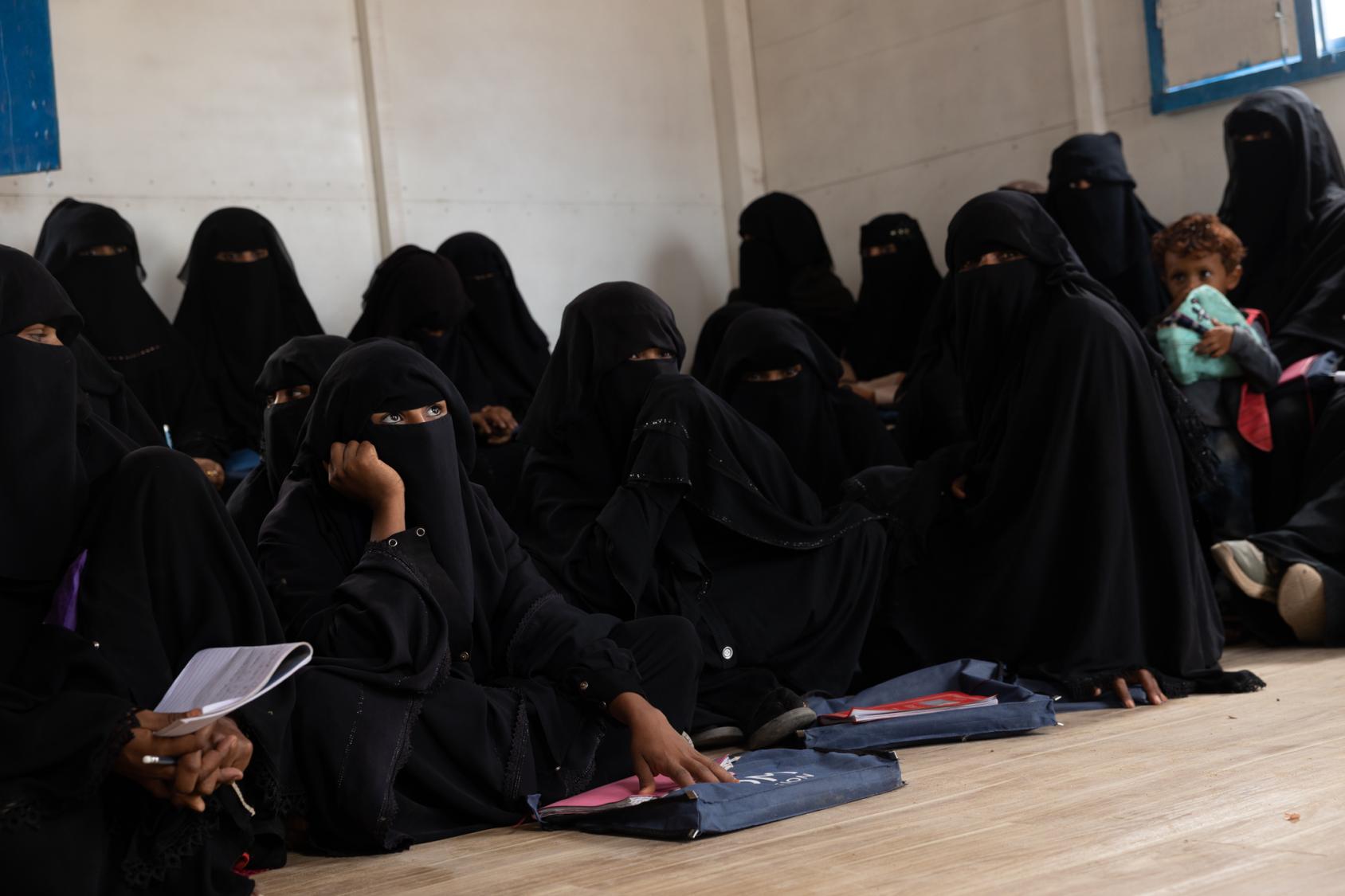 A row of women in full black clothing, sit on the floor with books laid out in front of them.