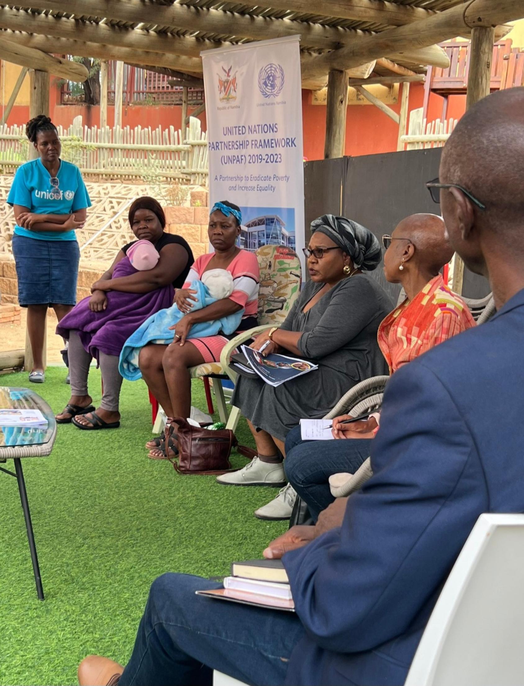 A group of people sit on chairs on a green carpet, talking to one another.