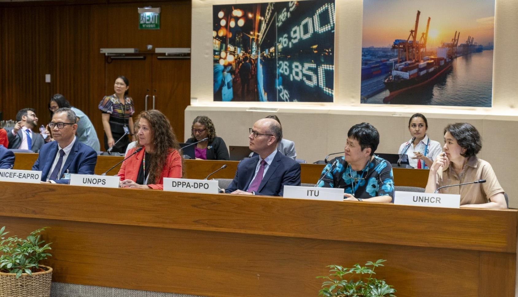 UN officials sit at a roundtable, with name tags and microphones, as they appear to listen.