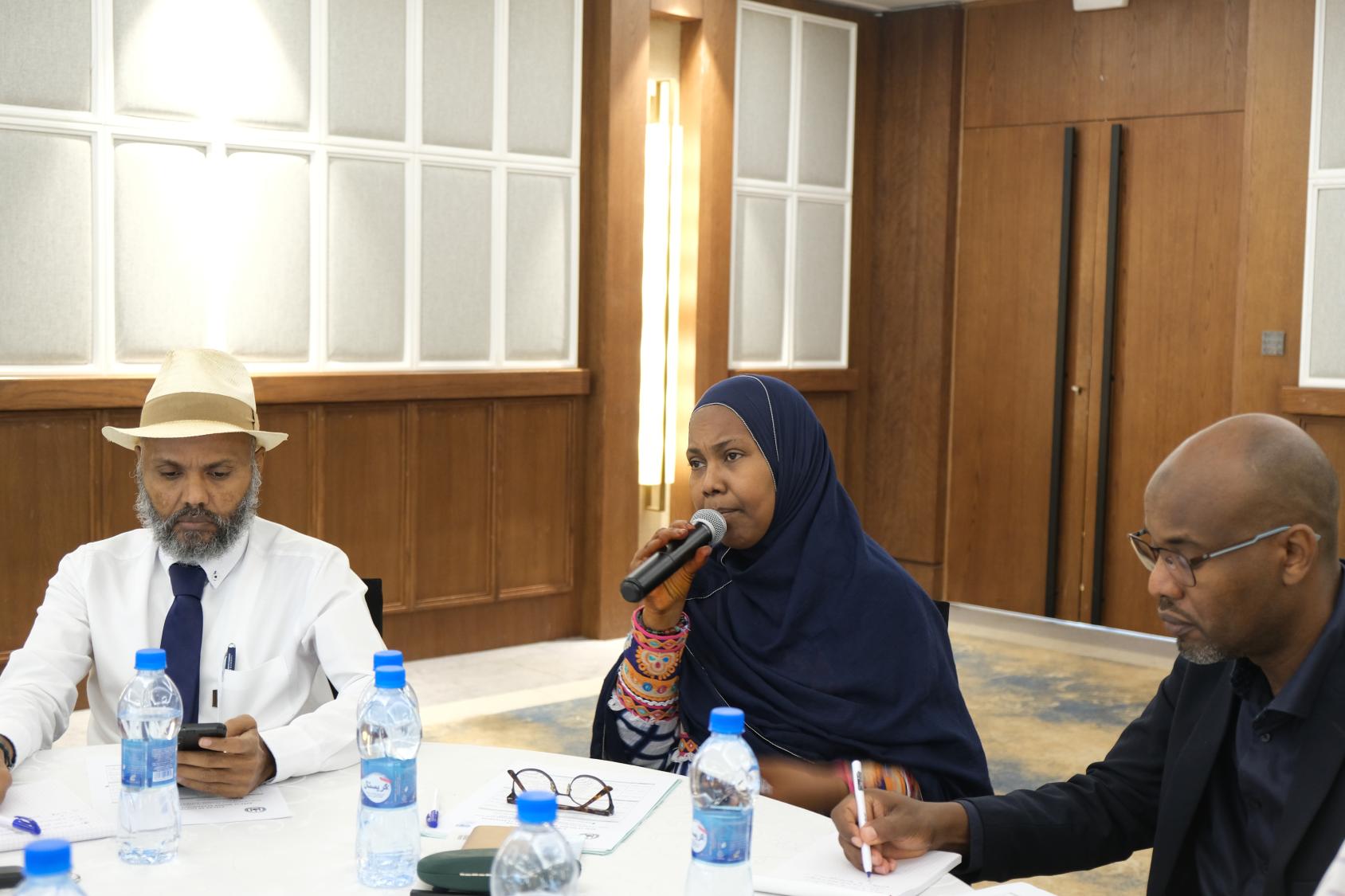 A woman in a black dress and headscarf sits on a table with two men on either side. She speaks into a microphone