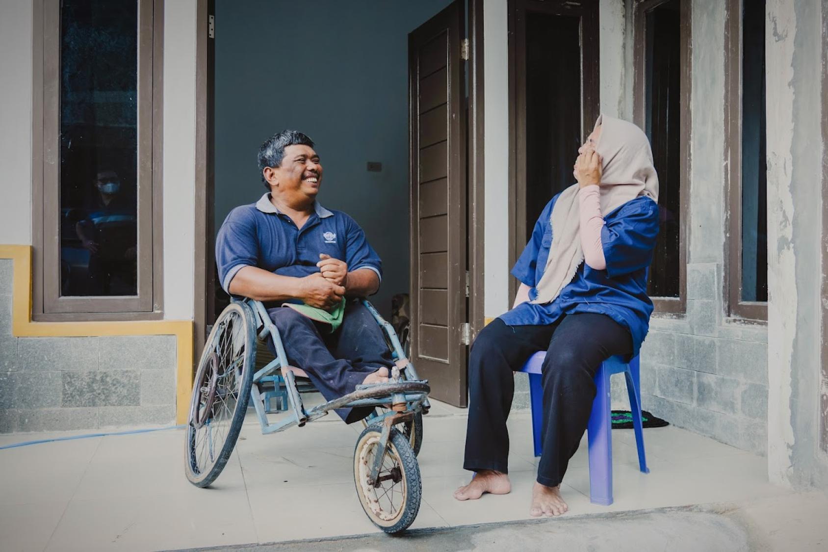 A person in a wheelchair and another seated on a stool engage in conversation outside a building with grey walls and brown doors. 