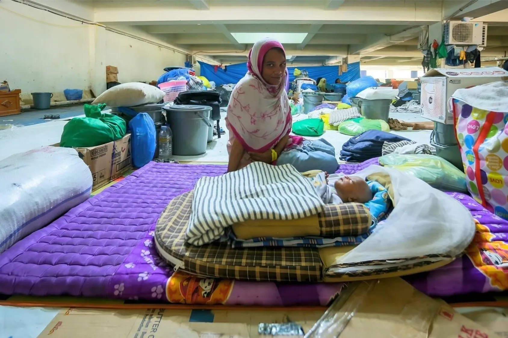 A woman in a dress and headscarf sits in a basement next to her sleeping child