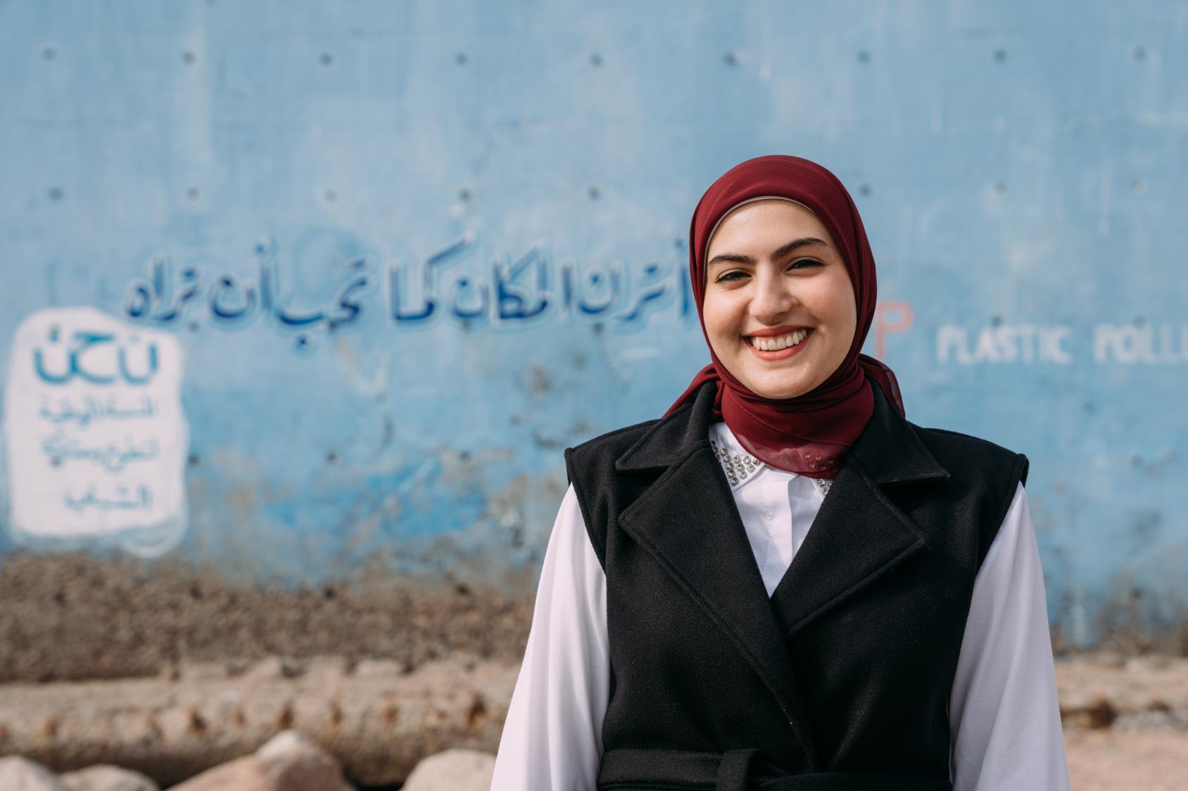 A woman in a black vest, white shirt and red headscarf stands in front of a blue wall with lettering on it