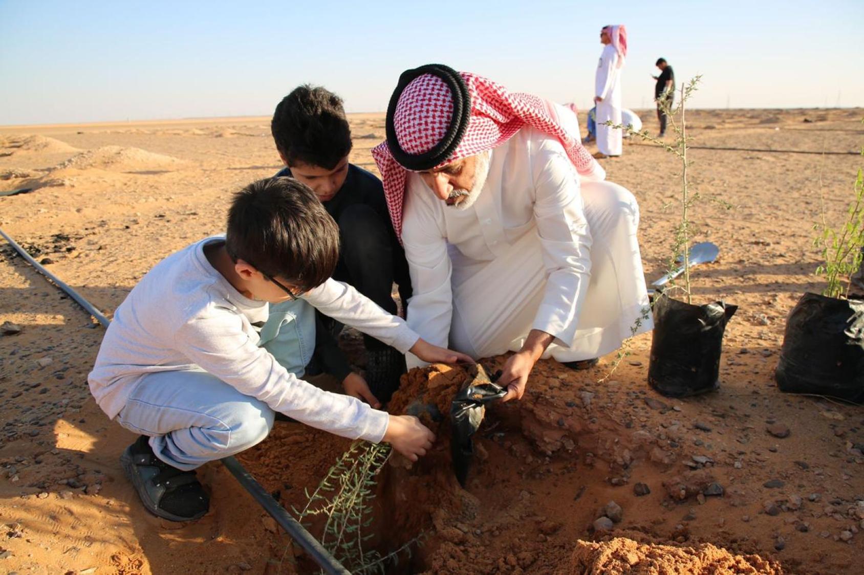 A man in a white dress and red headscarf plants a little shoot with two small boys.