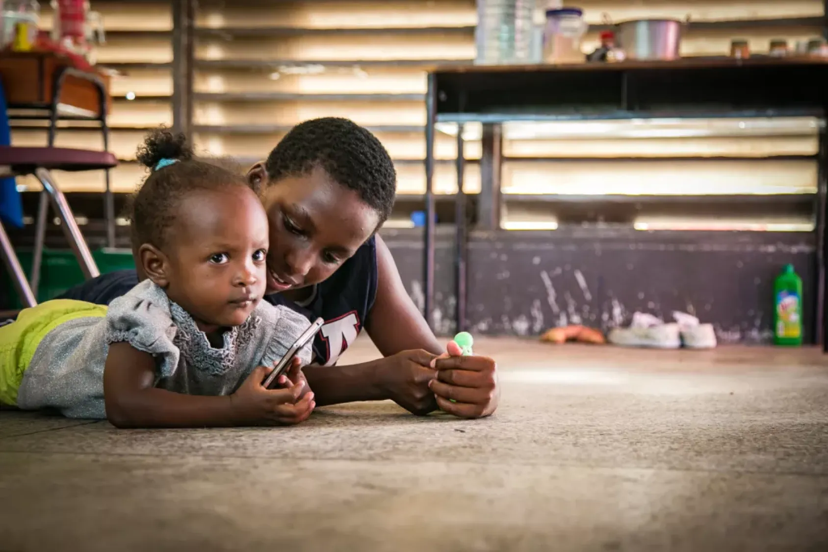 A young boy and a child lie on the ground cuddling next to each other. The child holds a cell phone in its hand.