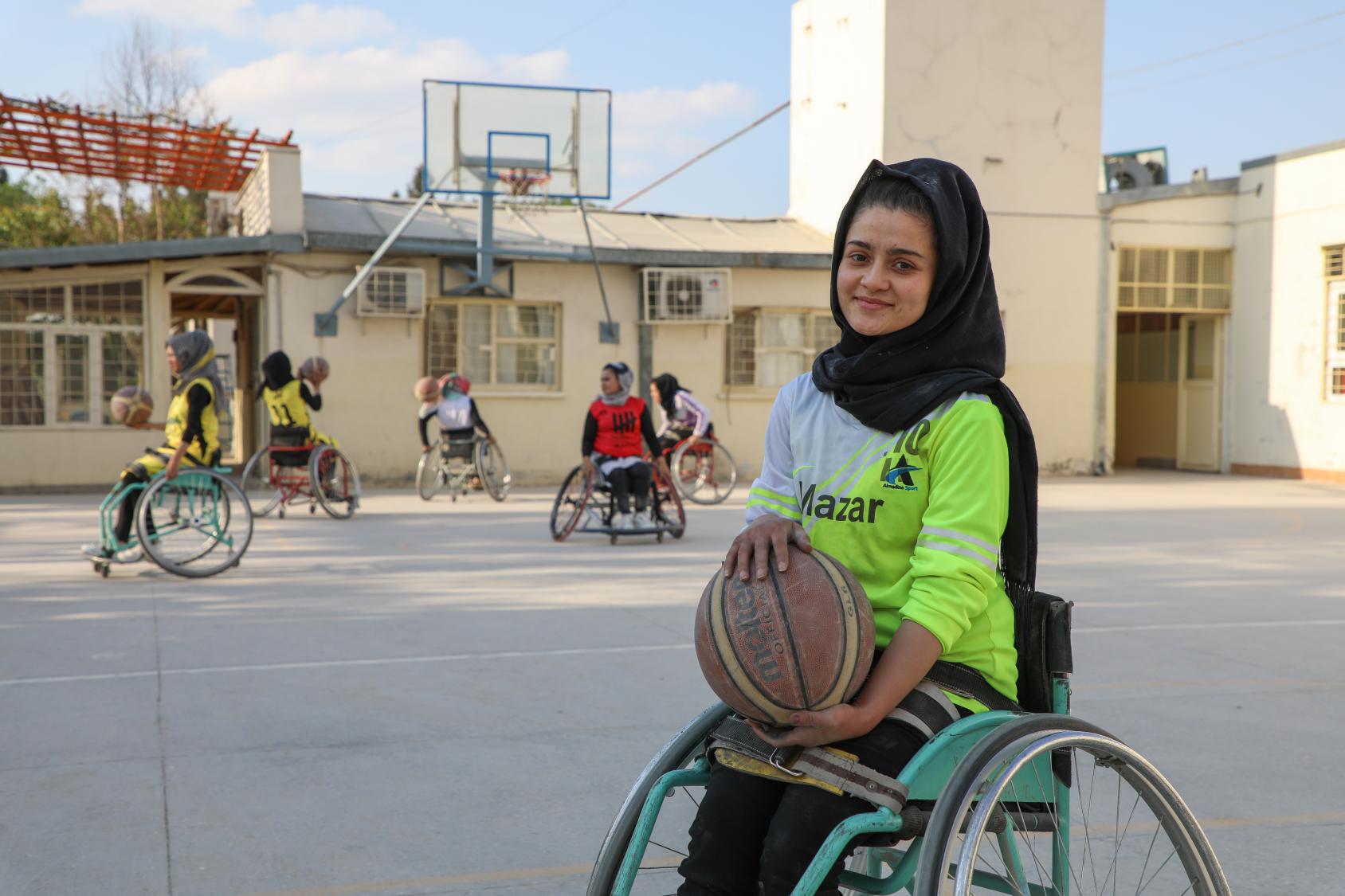 A girl in a neon green school uniform and black headscarf, with a ball in a wheelchair