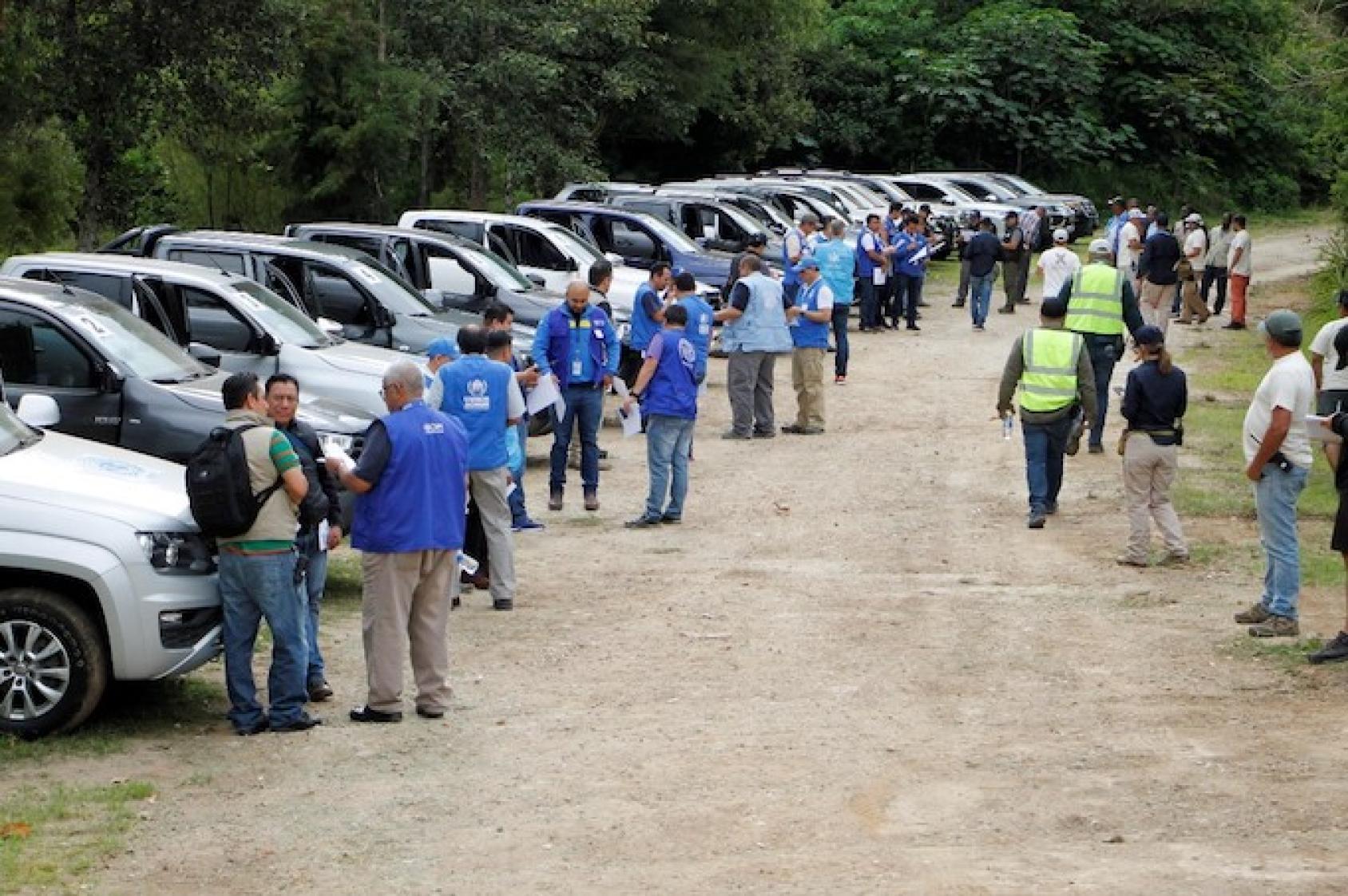 A line of cars outdoors with people in blue jackets standing by them. 