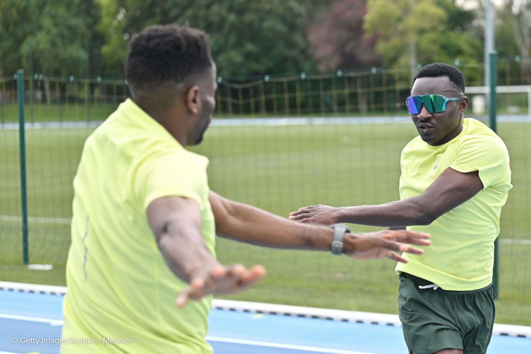 Two men in fluorescent green shirts, one in a dark visor, exercise on a running track.