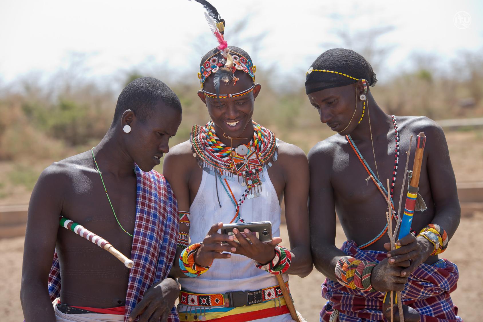 A group of three indigenous Kenyan men in traditional clothing look into a mobile phone