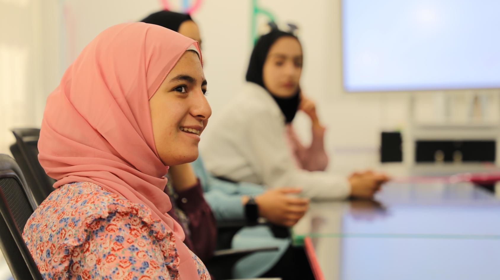 A girl in a pink scarf and top is looking to the side of the camera 