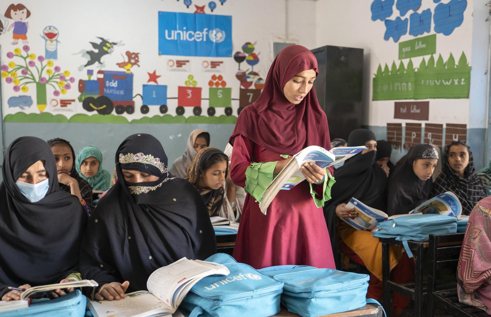 A girl in a red dress with her head covered reads from a book in a classroom. She stands amid other students who are seated in chairs.