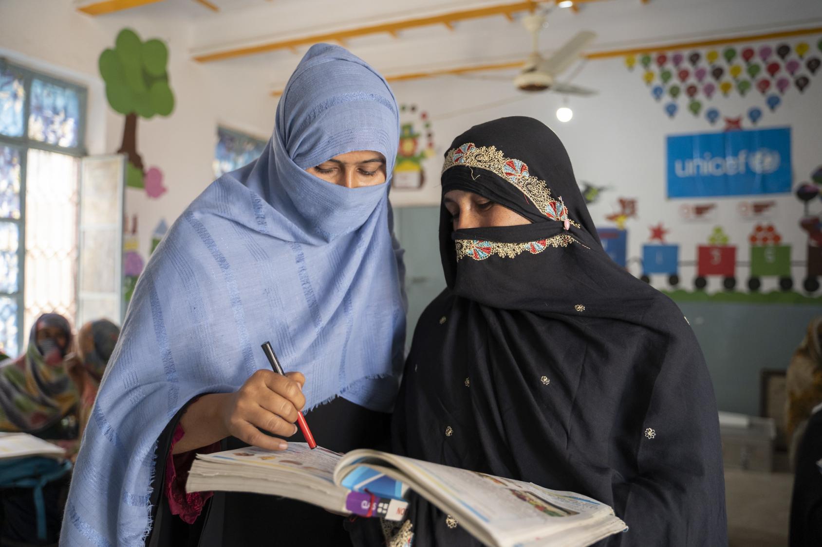 A woman in a black dress fully covered gets help from a teacher dressed in blue, with her face and head covered, over a notebook in a classroom with colourful walls.