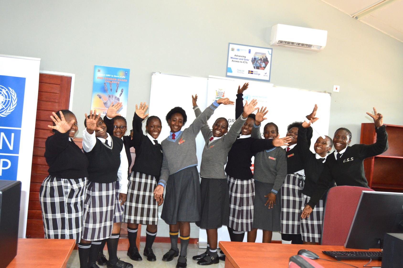 A group of eleven schoolgirls in uniforms wearing black and grey put their hands up joyfully in a classroom