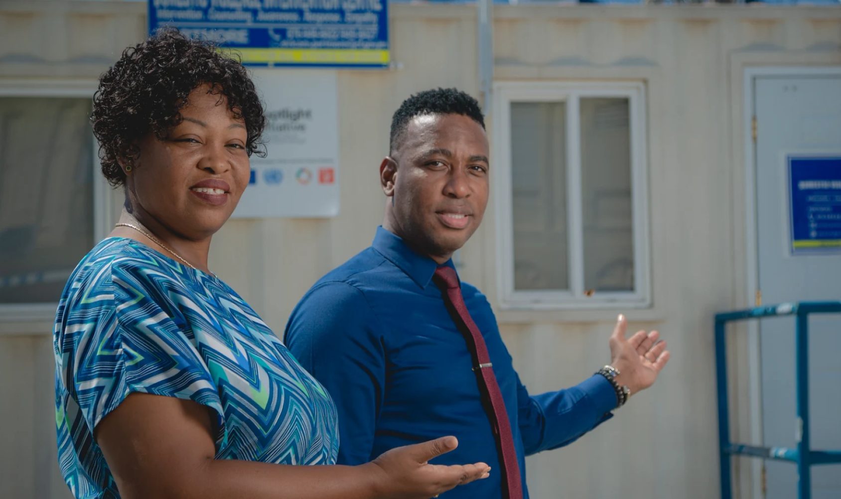 Two people, a woman and a man in blue uniforms and clothing point to a building entrance.