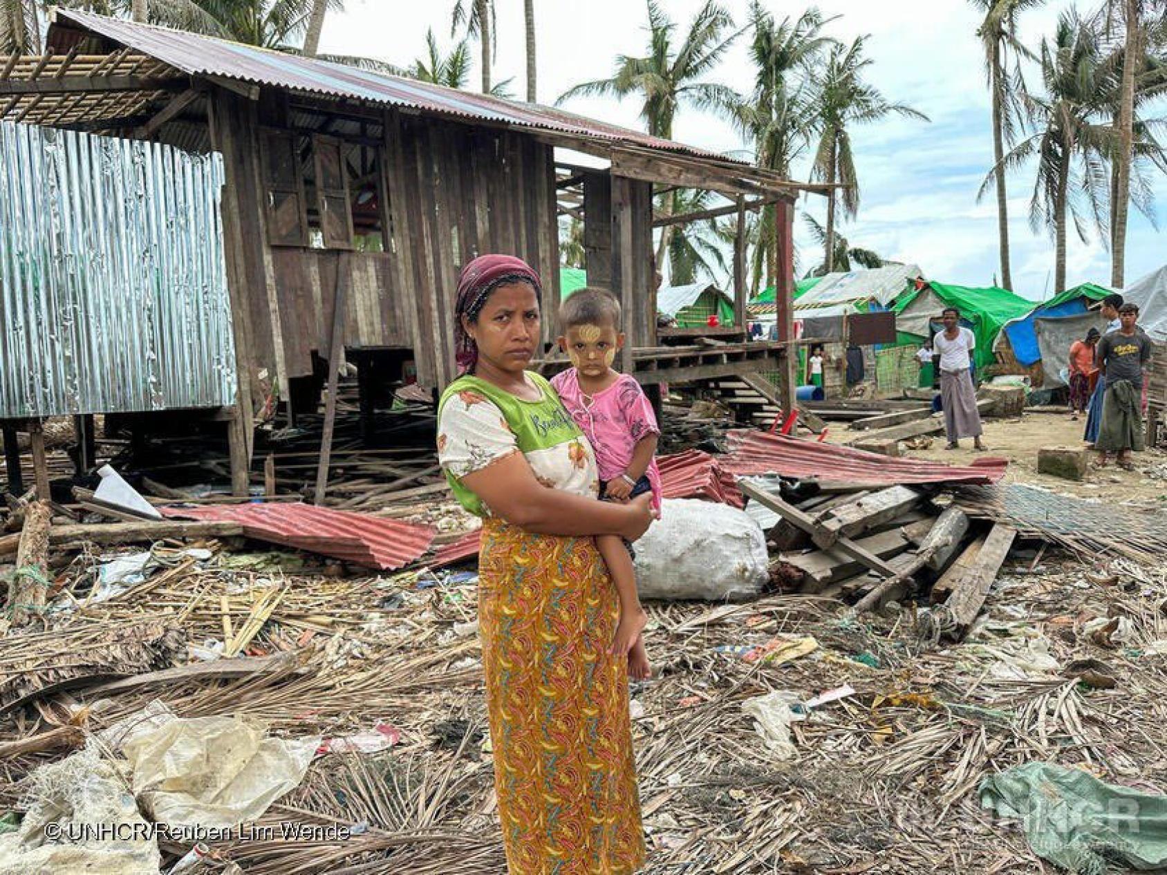 A woman in an orange skirt holds a baby in a pink dress amid rubble and a torn down house in the background