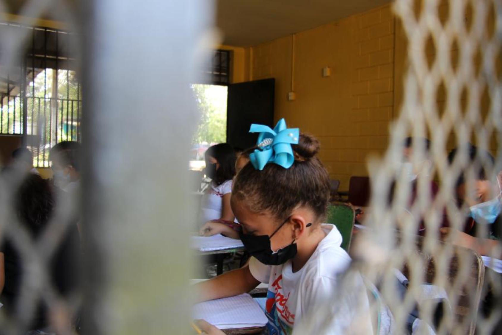 A young girl with a black mask on her face and blue bows studies inside a classroom in Honduras