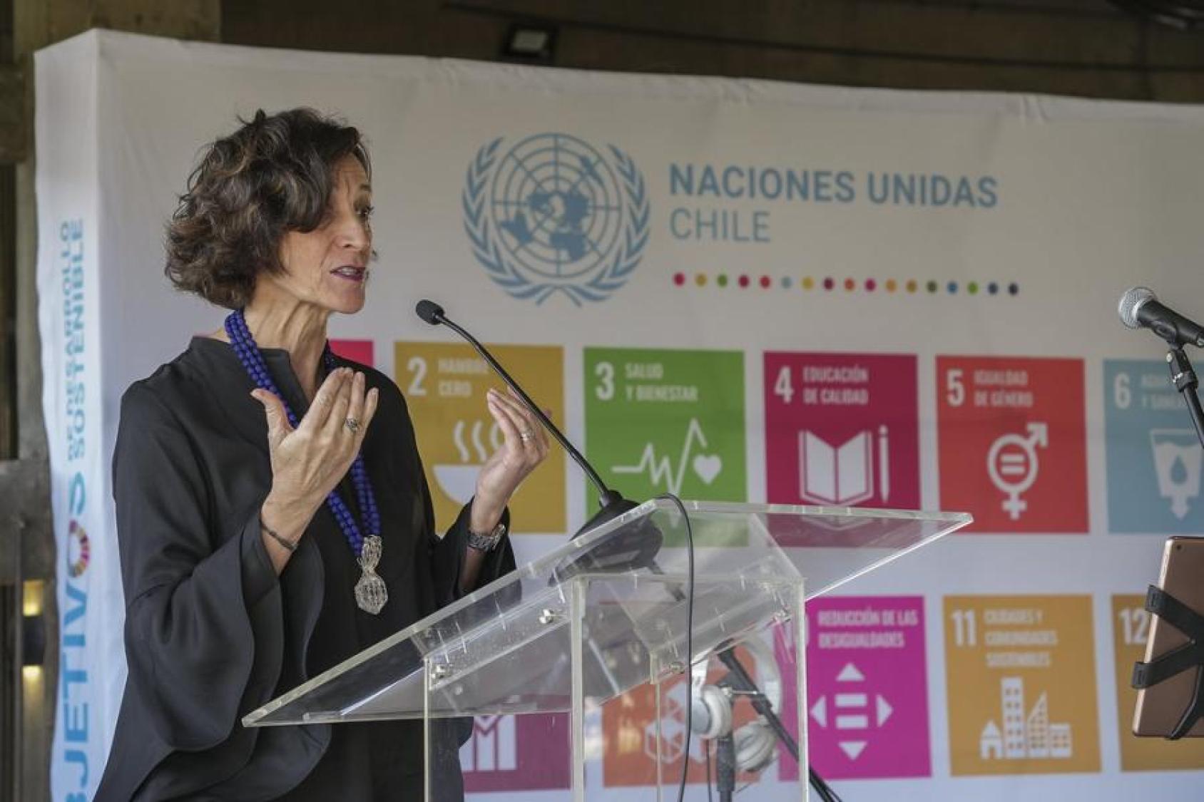A woman in a black dress speaks into a microphone at a podium. Behind her is a banner with the SDGs logos