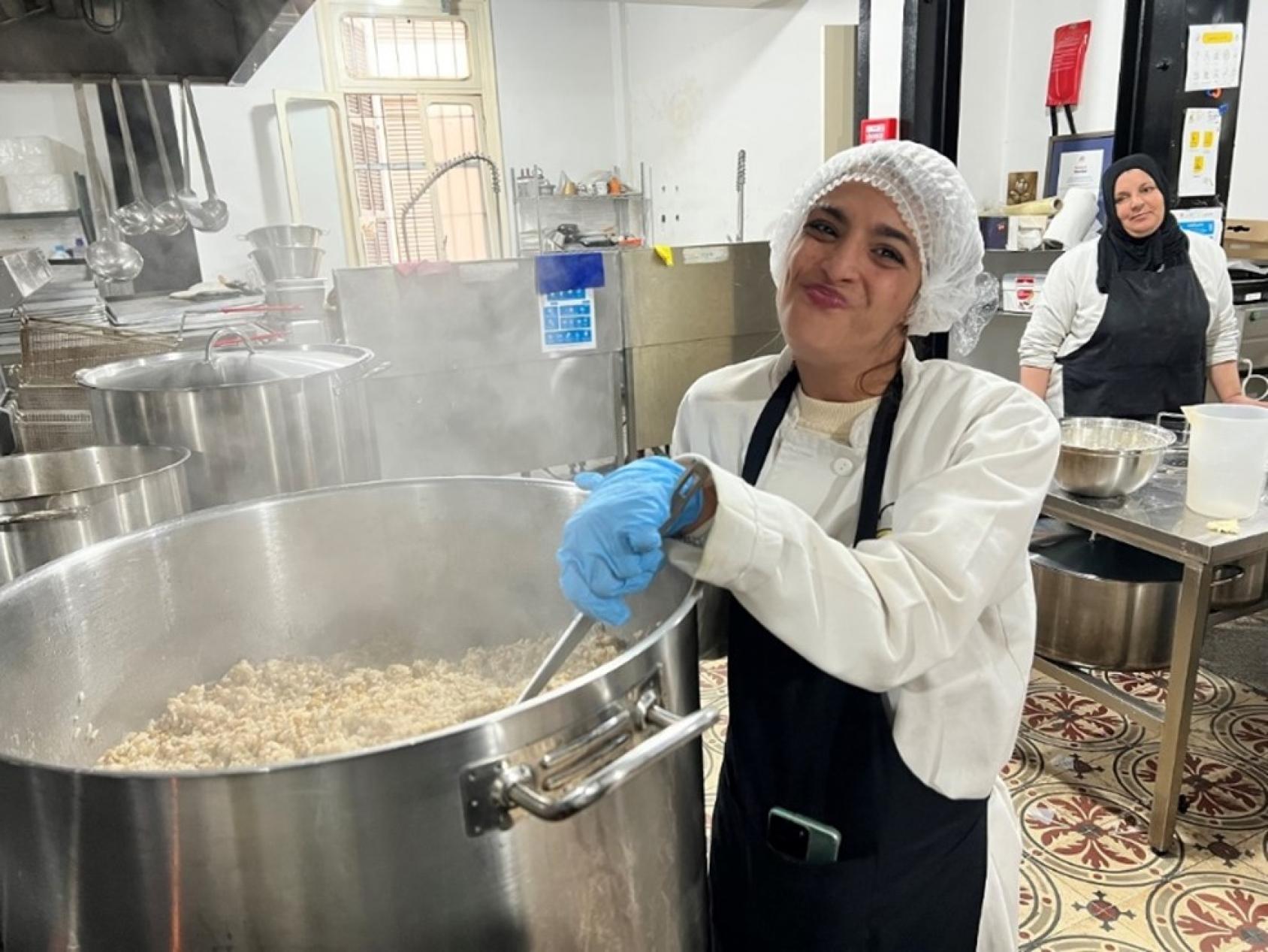 A woman in a white dress, black apron with blue gloves stirs food in a giant vessel in a kitchen