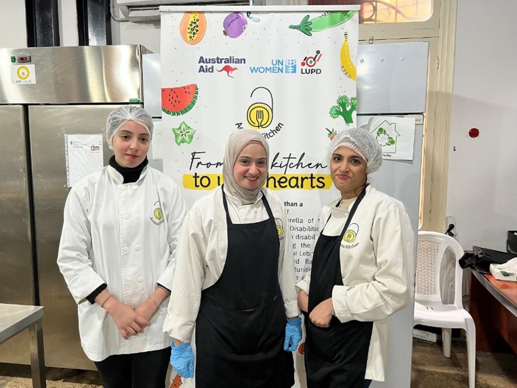 Three women in black aprons stand in front of a refrigerator and shelves inside a kitchen