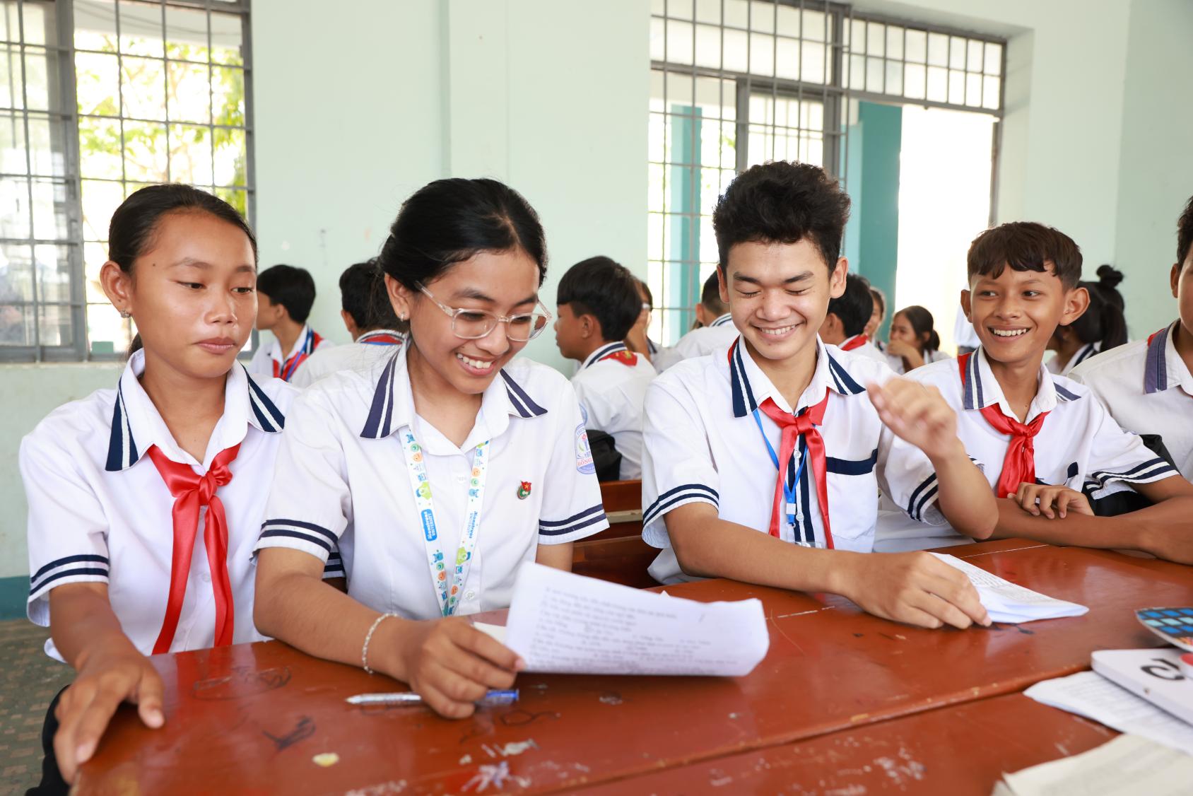 A classroom scene where two girl students sit next to two boy students in uniforms, as they all seem to be laughing.