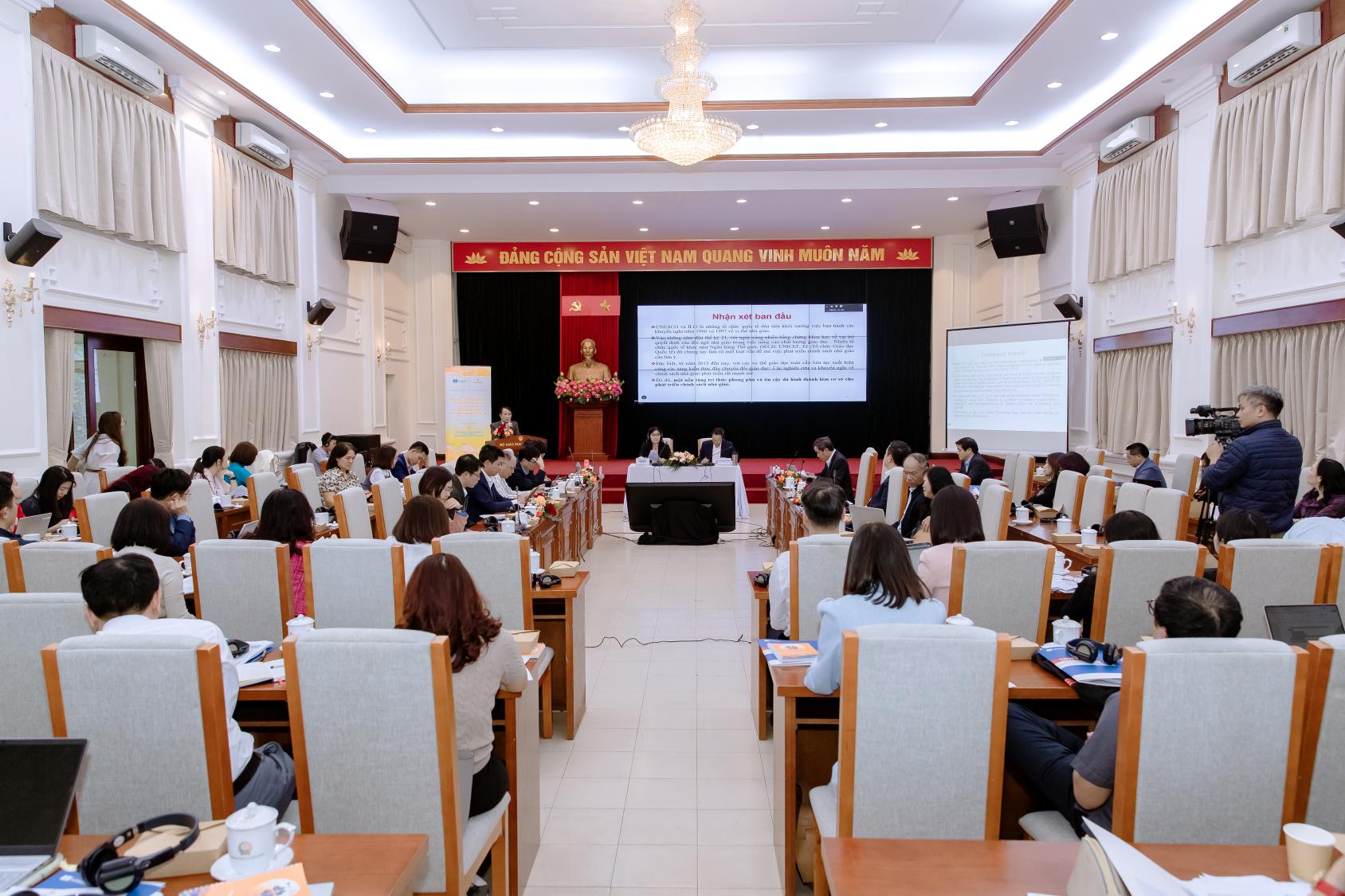 A hall where numerous people, teachers and experts, sit on white chairs and seem to be listening to a speaker at a podium.